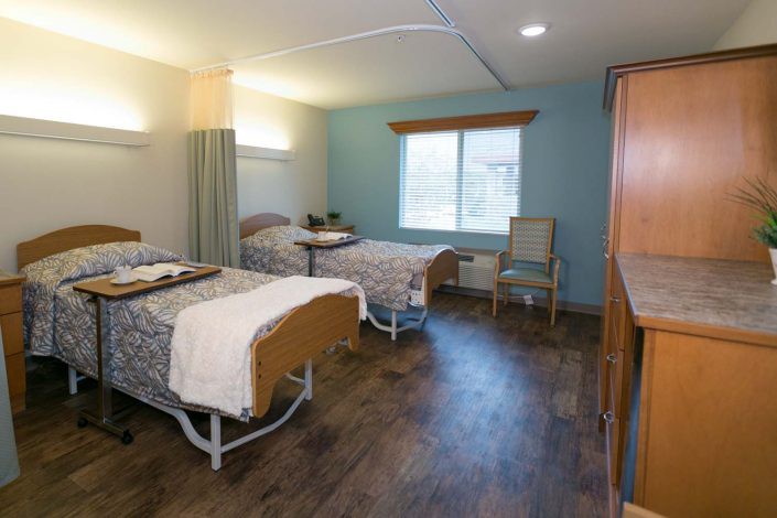 A hospital room with two patient beds separated by a curtain, featuring wooden flooring and a window providing natural light.