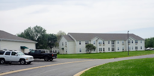 A two-story residential building surrounded by grass and trees, with a truck driving along the curved road.