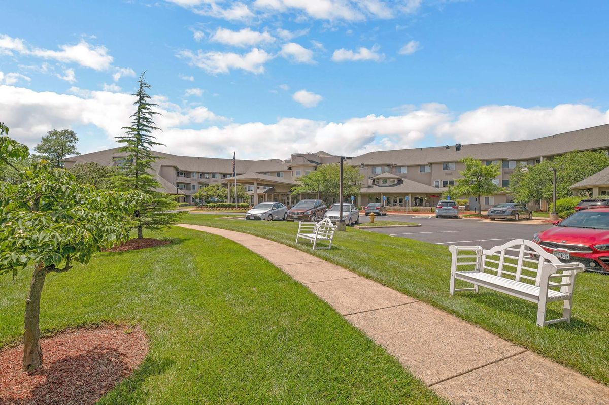A view of a large building with a neatly maintained lawn, trees, and benches, adjacent to a parking lot.