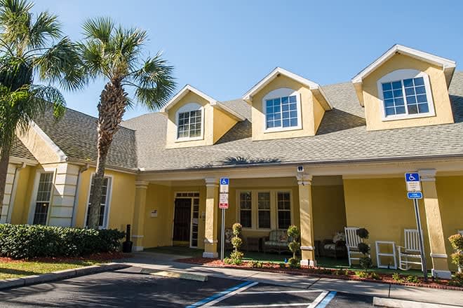 A yellow building with a pitched roof and multiple windows, featuring a palm tree in front and an adjacent parking lot.