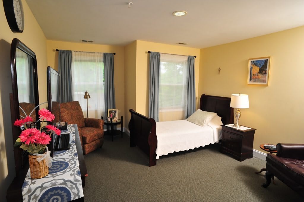 A cozy bedroom featuring a single bed, armchair, and nightstand with a lamp, complemented by two windows with blue curtains.