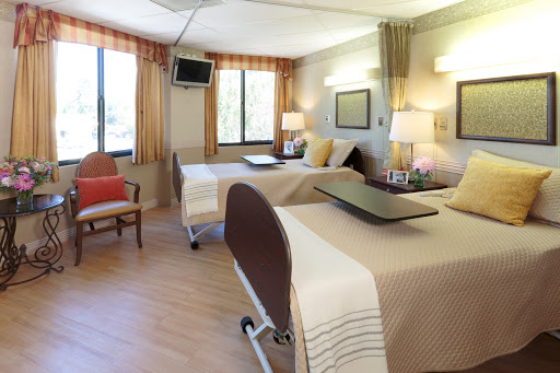 A spacious and well-lit hospital patient room with two beds, natural light from windows, and comfortable furnishings.