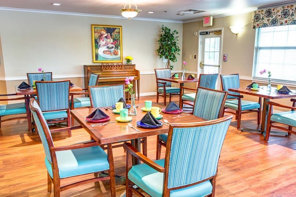 A well-decorated dining room featuring neatly arranged tables and chairs, with a piano and artwork in the background.
