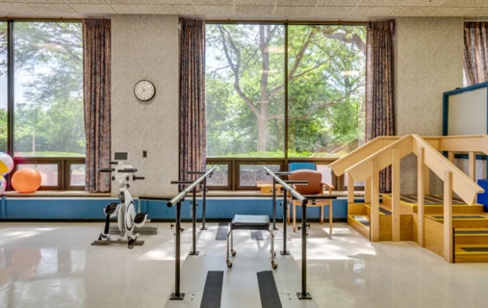 A physical therapy room equipped with parallel bars, an exercise bike, and a small staircase, providing a bright and inviting setting for rehabilitation exercises.