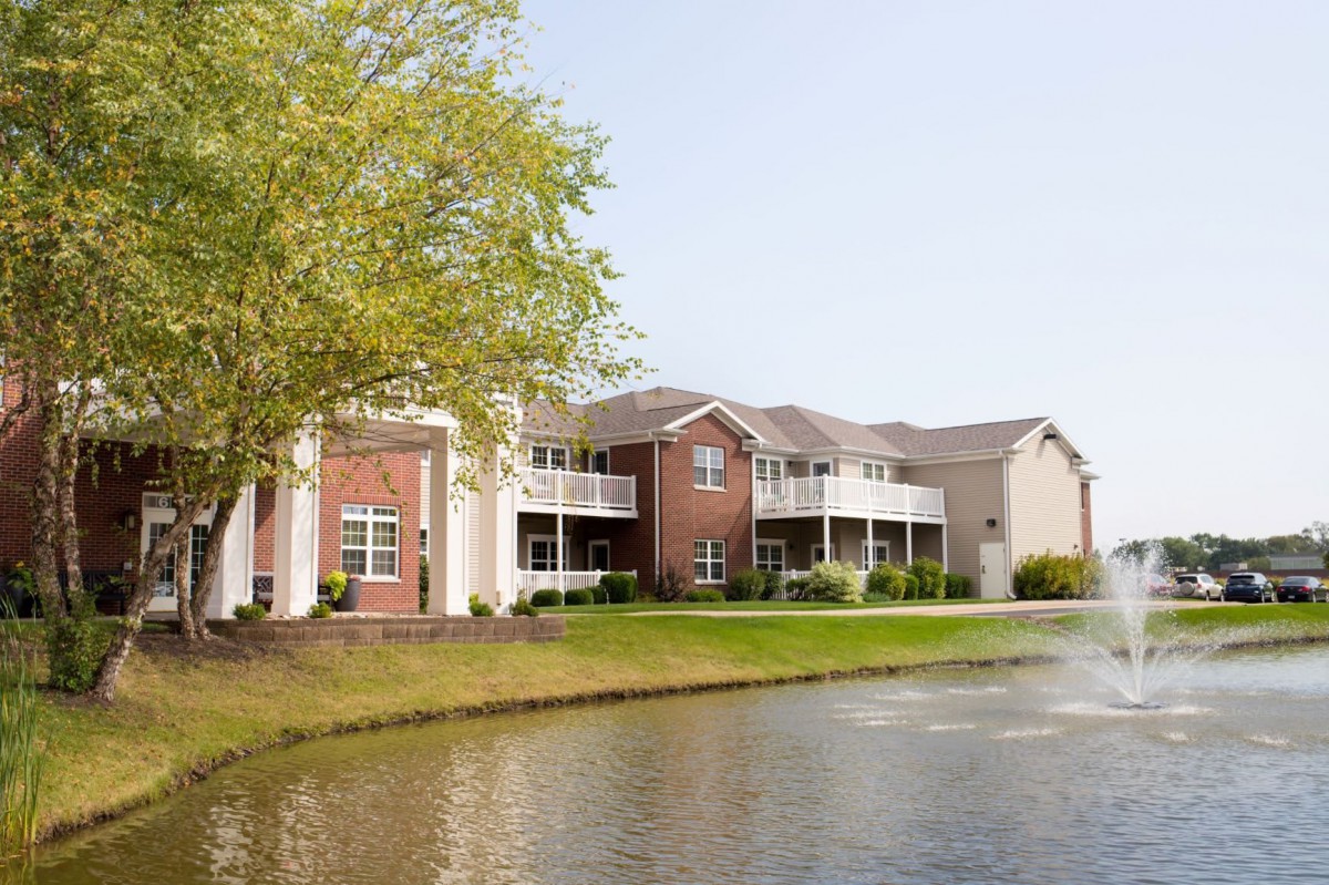 A scenic view of an apartment building by a tranquil lake, surrounded by greenery.