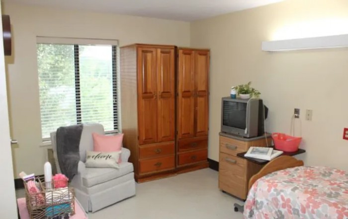 A cozy bedroom with a wooden wardrobe, comfortable chair, and an old-style TV near the window.