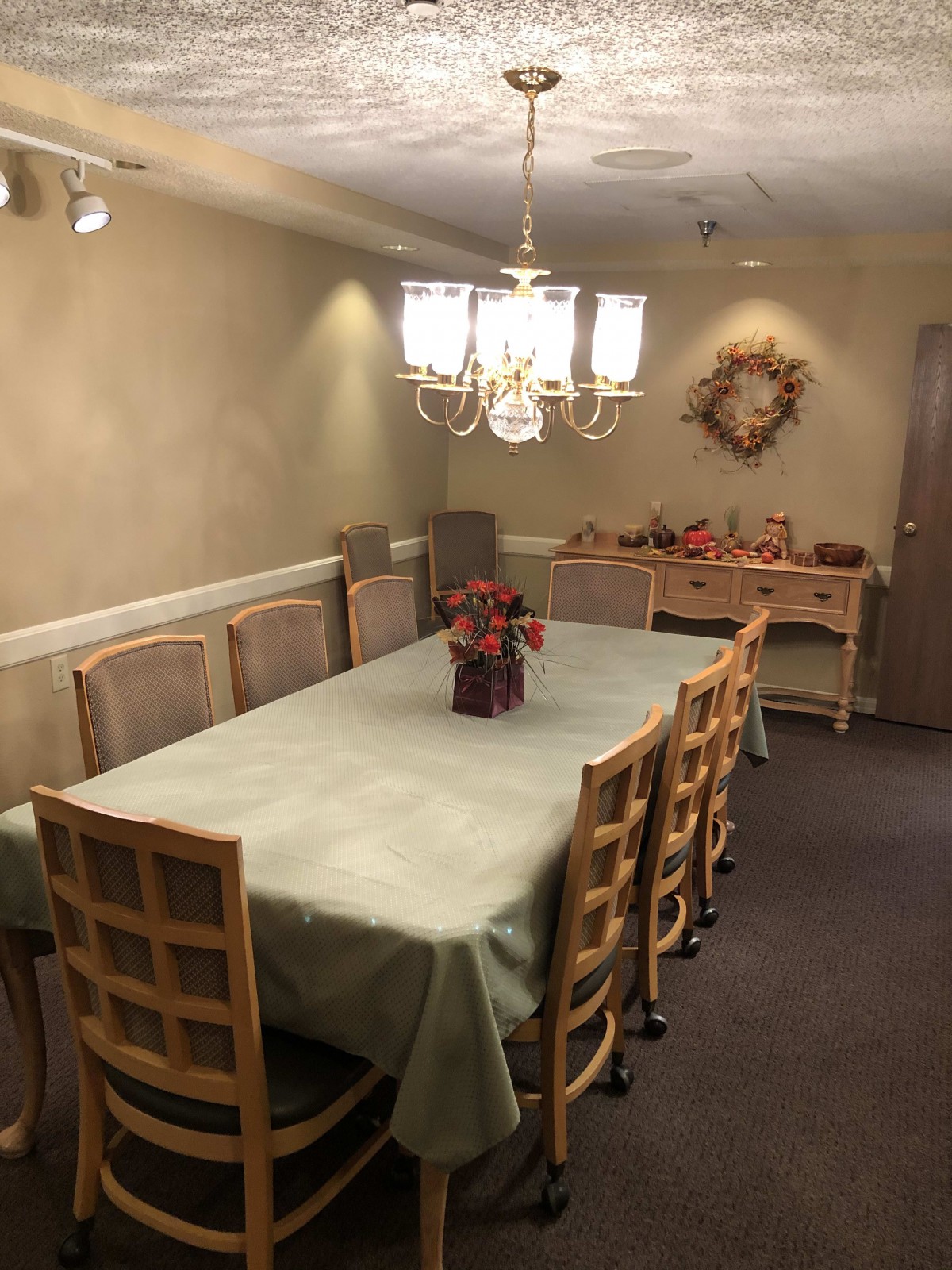 A cozy dining room featuring a long table covered with a tablecloth, surrounded by chairs, and illuminated by a chandelier.