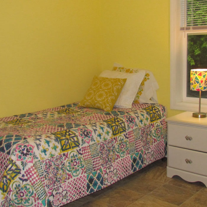 A cozy bedroom with yellow walls, featuring a single bed with a colorful patchwork bedspread and a nightstand with a lamp by the window.