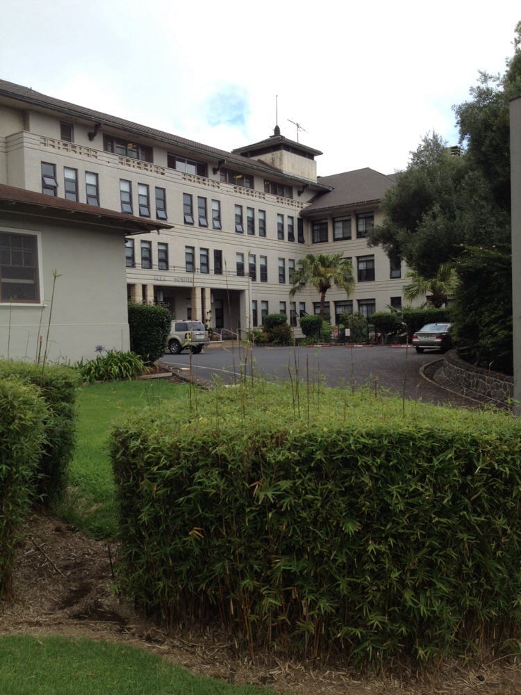 A view of a multi-story building with cars parked in the driveway and lush greenery surrounding the area.