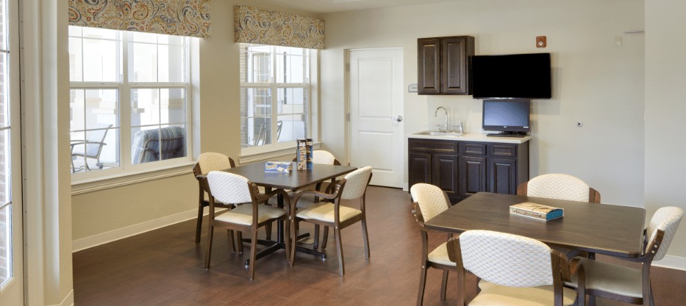 A well-lit dining area with a modern table and chairs, adjacent to a kitchenette with a television.