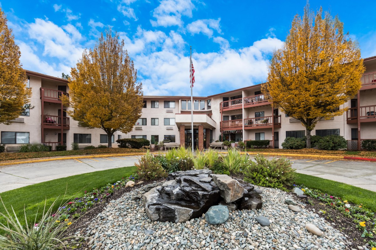 A multi-story residential building with autumn trees and a landscaped entrance under a partly cloudy sky.