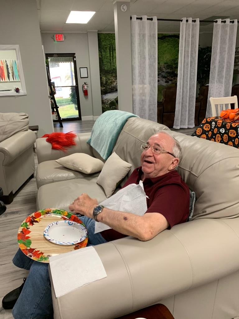 An elderly man sitting on a sofa in a living room, holding a plate and laughing.