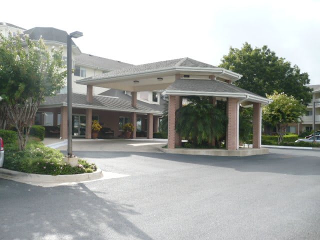 The image shows the entrance to a residential building with a covered driveway and surrounding greenery.