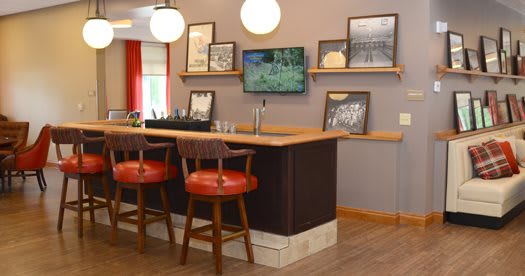 A cozy bar interior featuring a wooden counter, red cushioned chairs, framed pictures on the walls, and a small television.