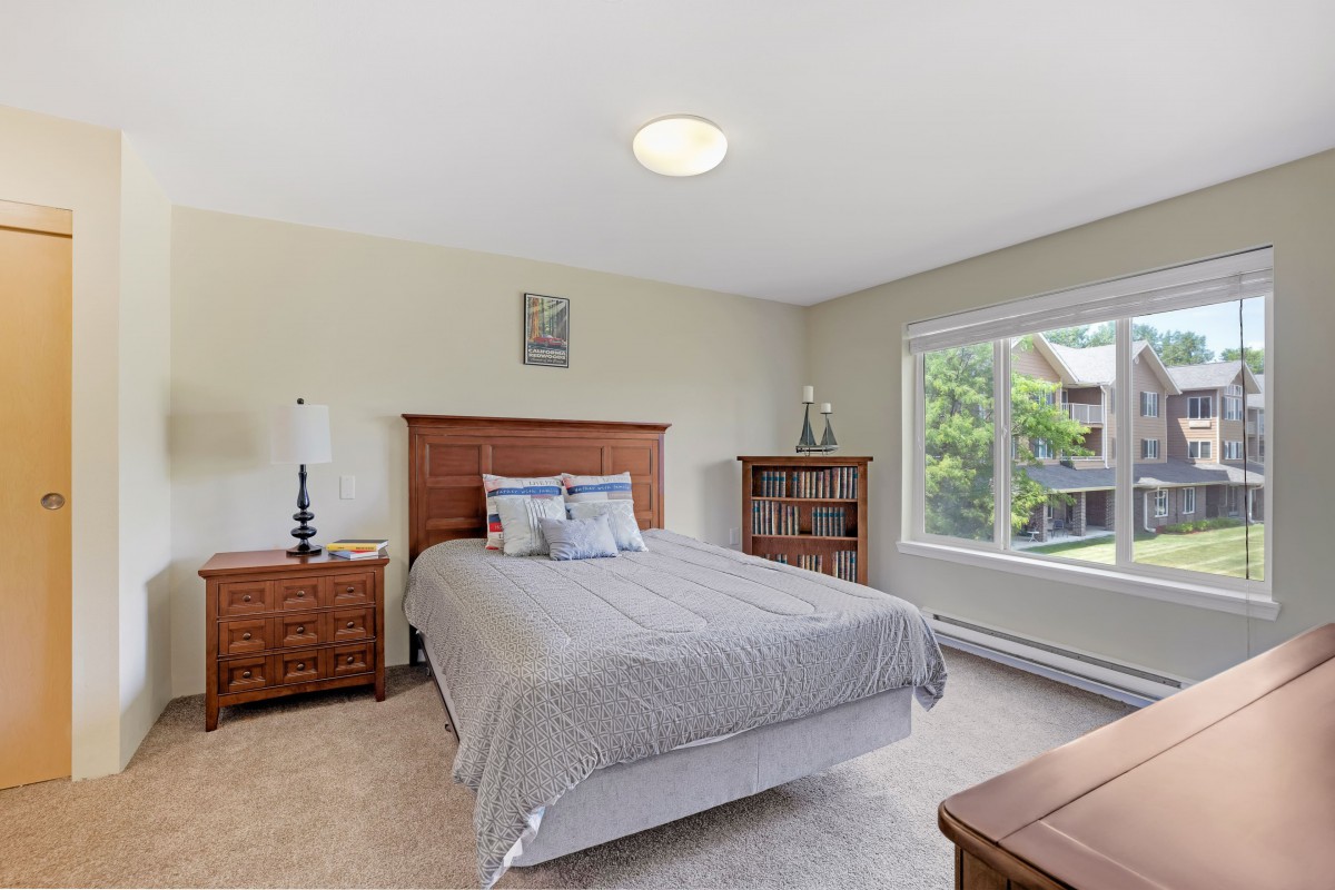 A cozy bedroom with a neatly made bed, wooden furniture, and a large window providing natural light.