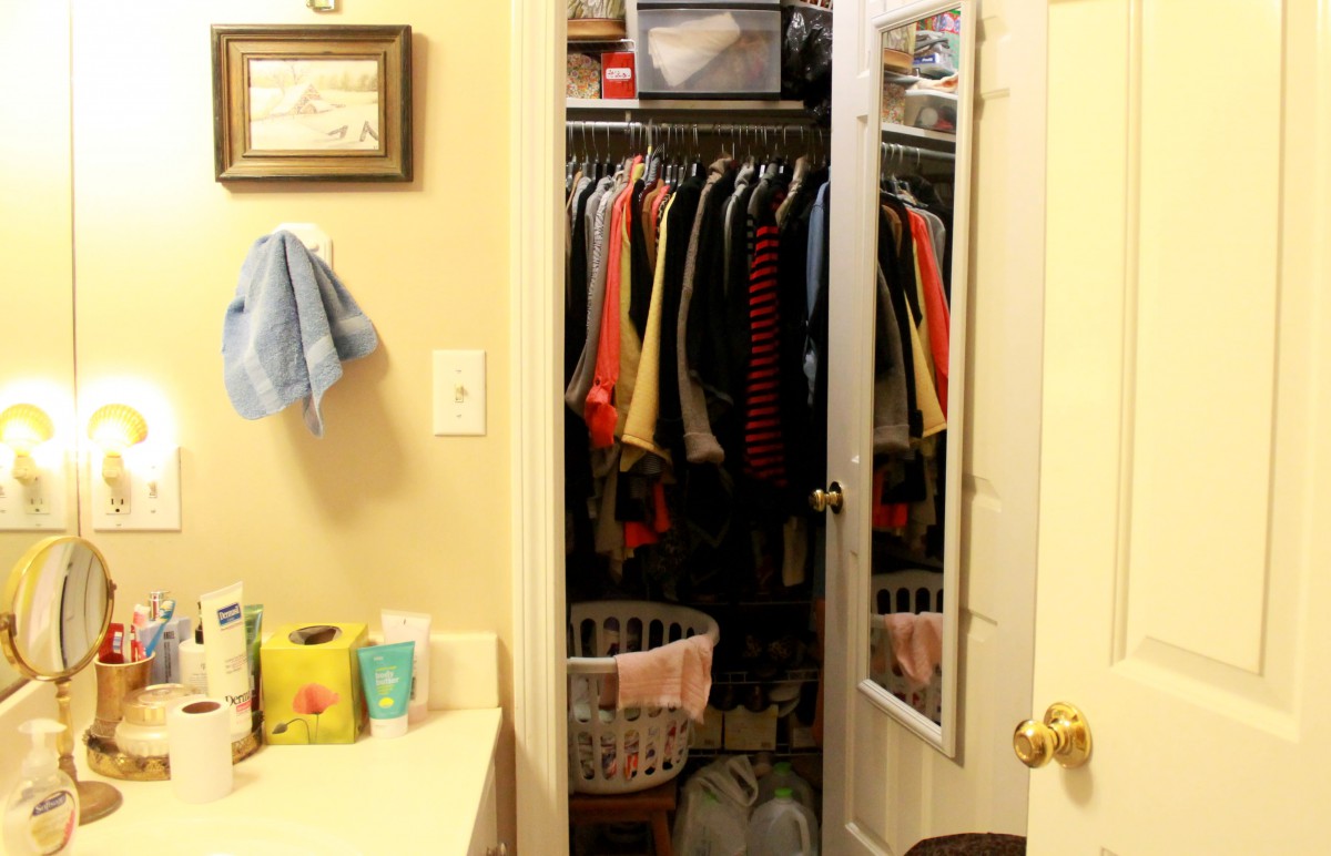 A bathroom with an open closet full of hanging clothes and a laundry basket.
