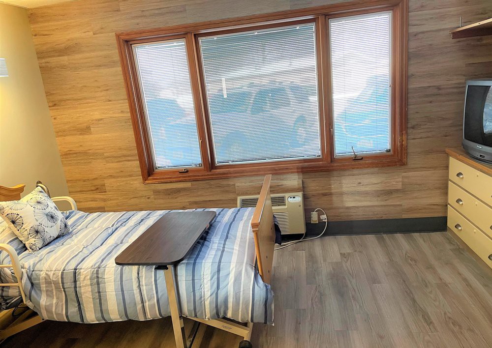 A simple room with wooden walls featuring a hospital bed by a large window with blinds.