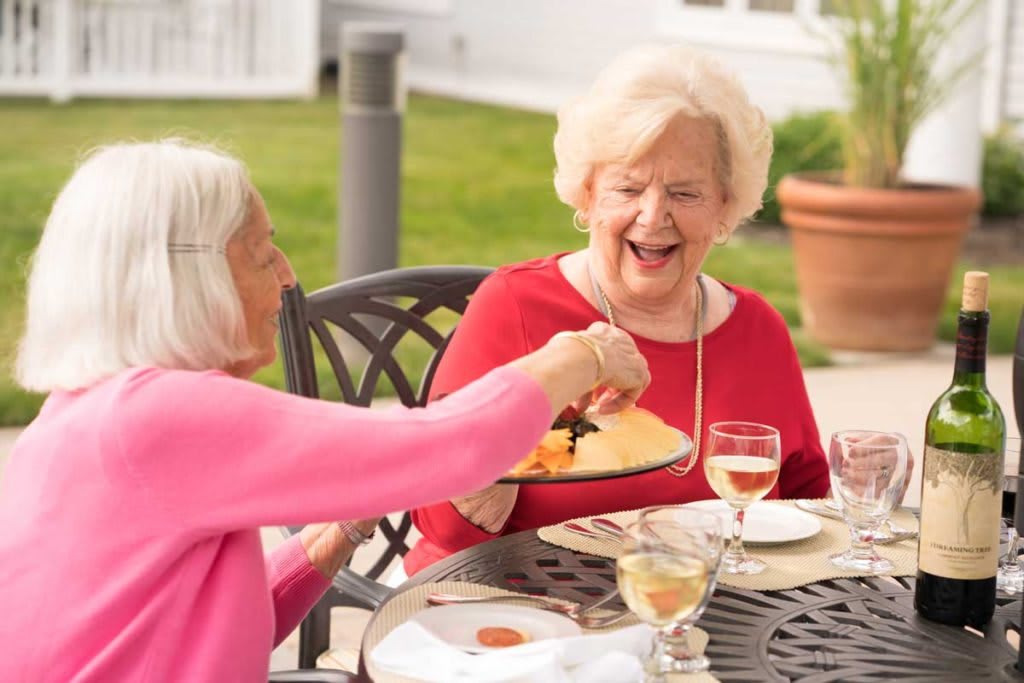 Two elderly women enjoying a meal and laughing together outdoors.