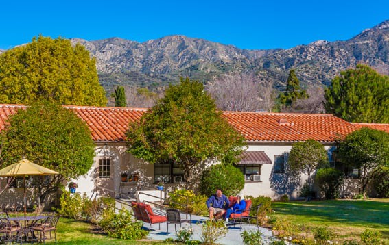 A charming home with a red-tiled roof set against a backdrop of mountains, featuring a lush garden and patio where people are relaxing.
