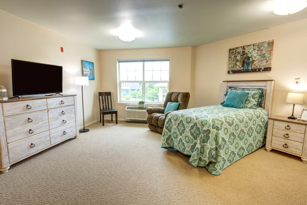 A cozy bedroom with a bed, chair, television, and a window providing ample natural light.