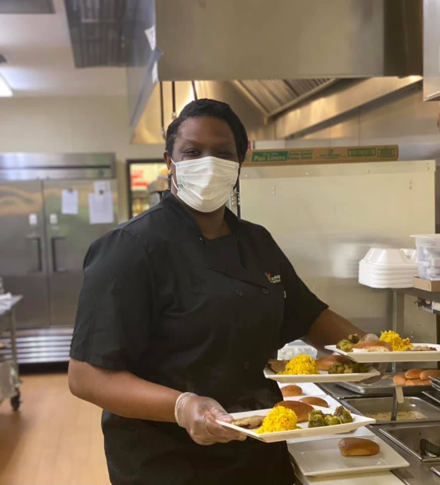 A chef in a commercial kitchen preparing plates of food while wearing a mask and gloves.