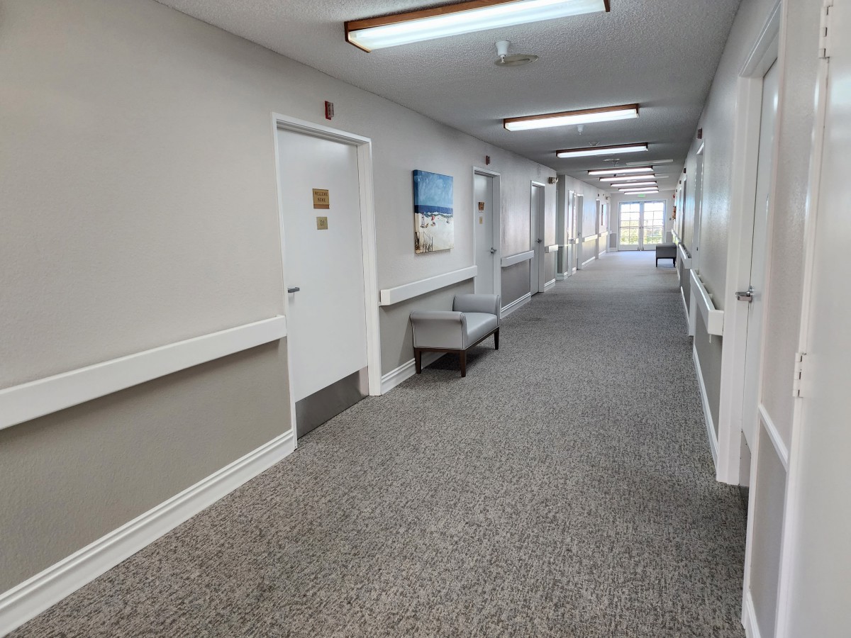 A long, carpeted hallway with several doors, ceiling lights, a bench, and paintings on the walls.