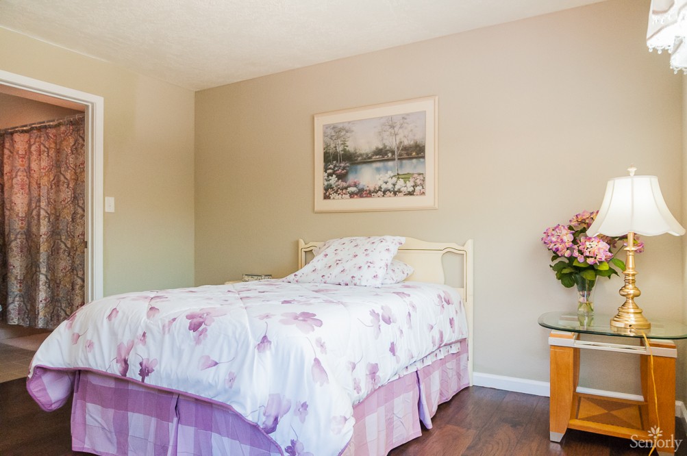 A cozy bedroom with floral bedding, a framed painting on the wall, and a nightstand with flowers.