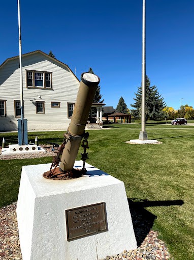 A small cannon monument on a pedestal in front of a building with flagpoles and green lawns under a clear blue sky.