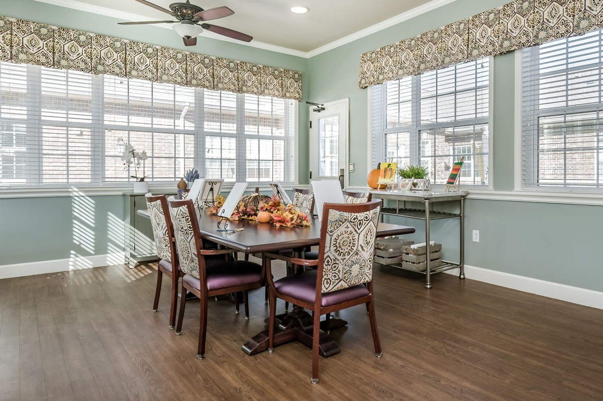 A well-lit dining room with a wooden table surrounded by upholstered chairs, featuring large windows and elegant decor.