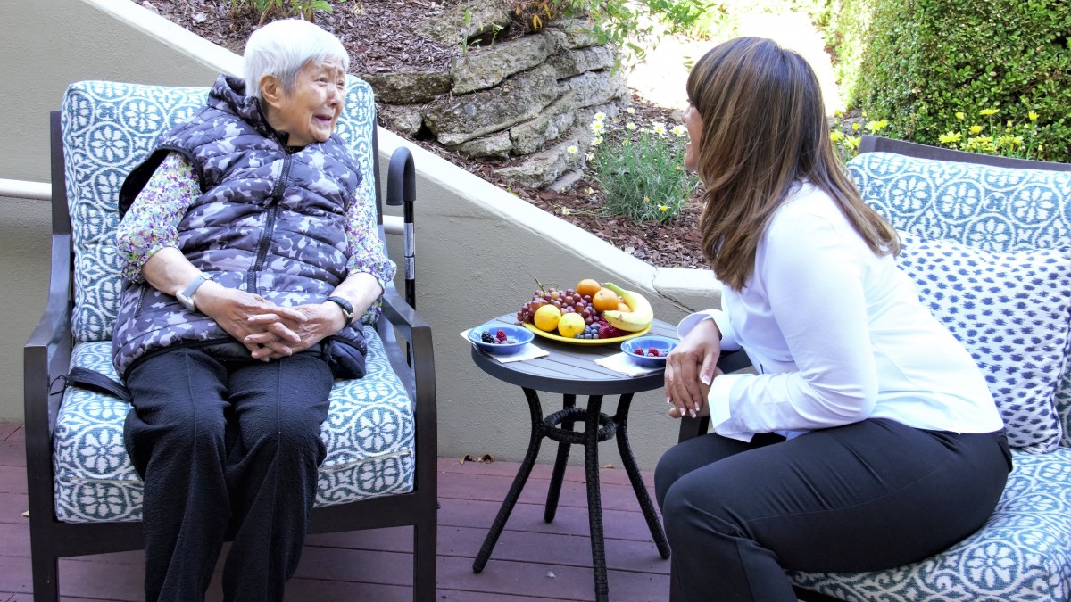 Two women, one elderly and one young, engage in a conversation on a patio with a table of fruits between them.