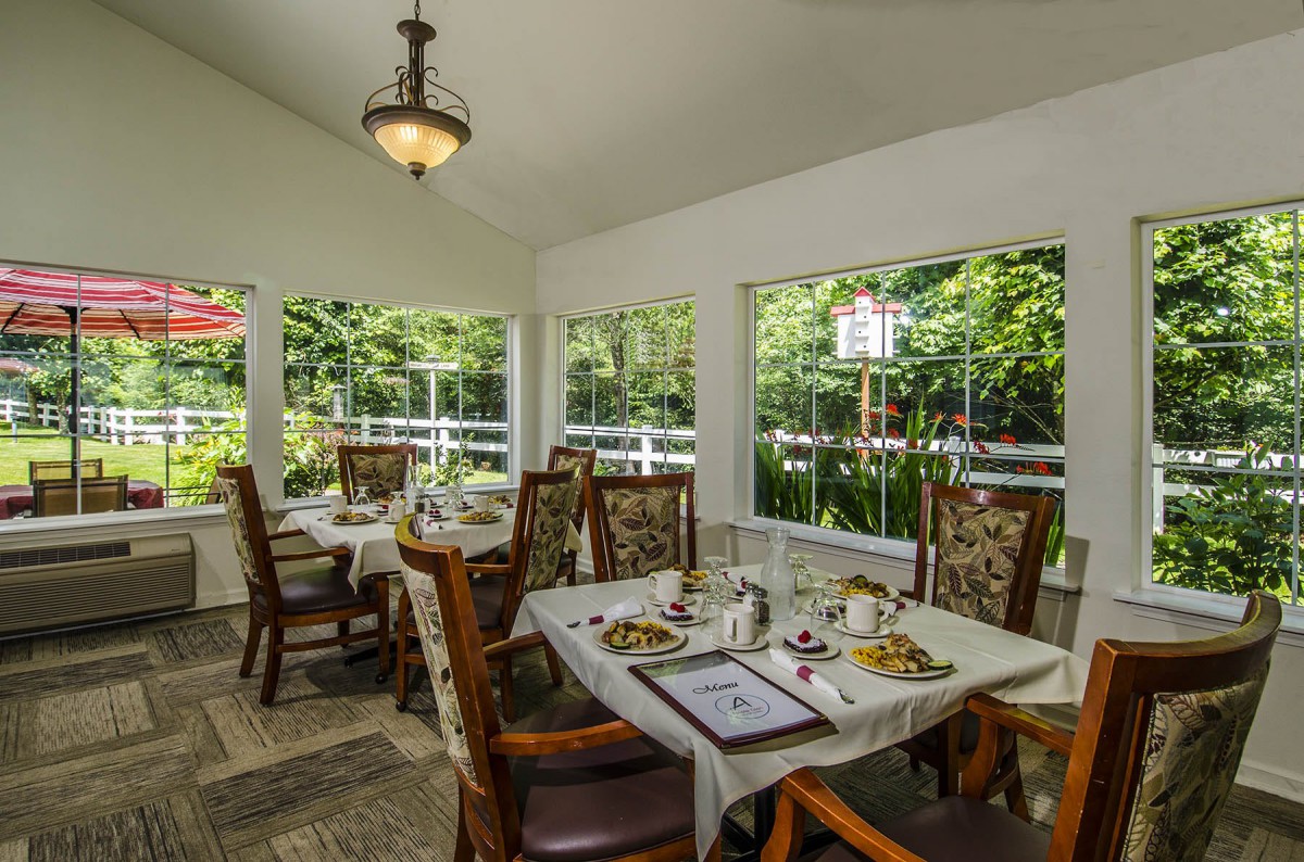 A cozy dining room with elegant tables and chairs, surrounded by large windows showcasing lush greenery.