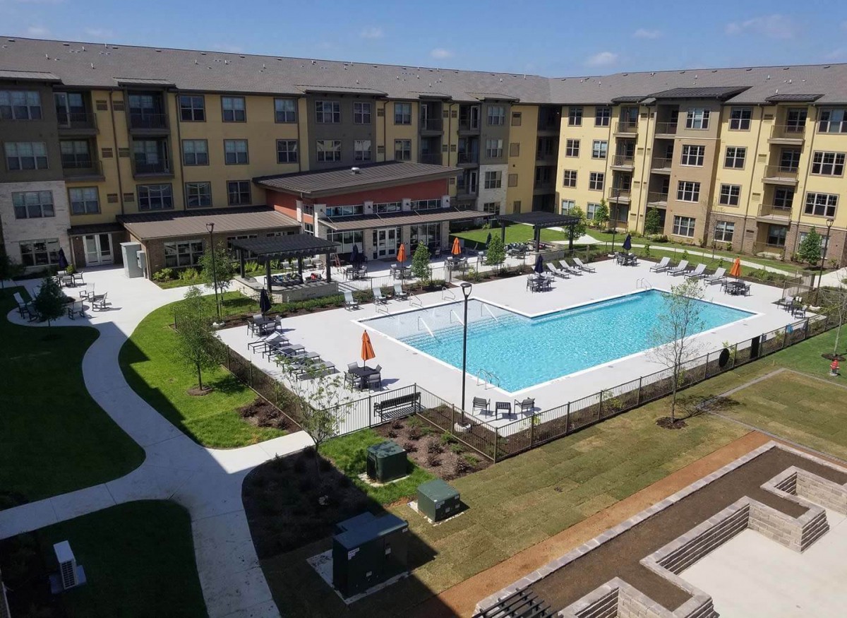 Aerial view of an apartment complex featuring a central swimming pool and landscaped outdoor area on a sunny day.