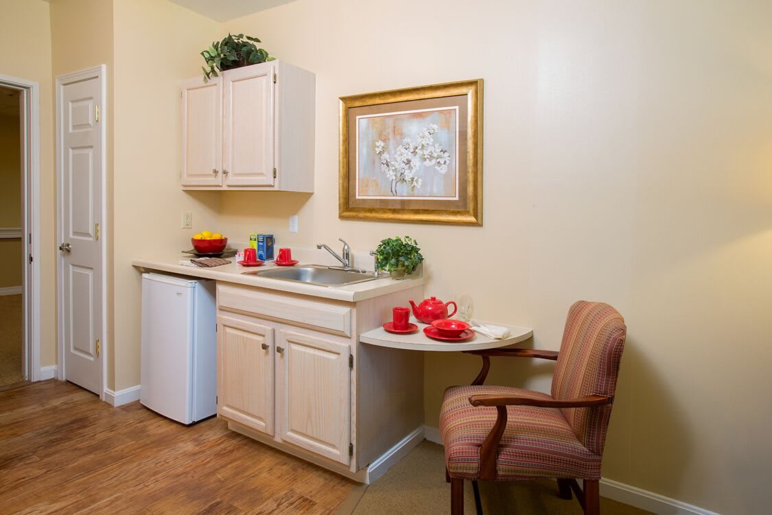 A cozy kitchenette with a small fridge, sink, and cabinets, featuring a table set for tea and decorative elements.