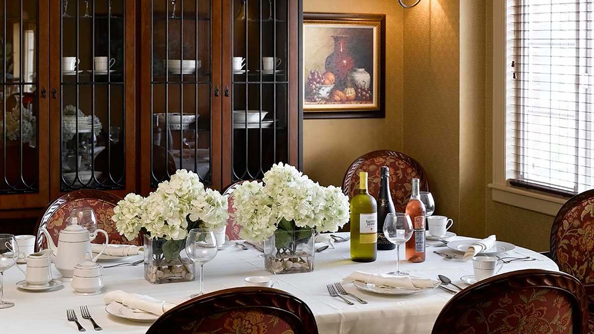 An elegantly set dining table with flowers and wine bottles, surrounded by ornate chairs, a decorative cabinet, and a framed painting on the wall.