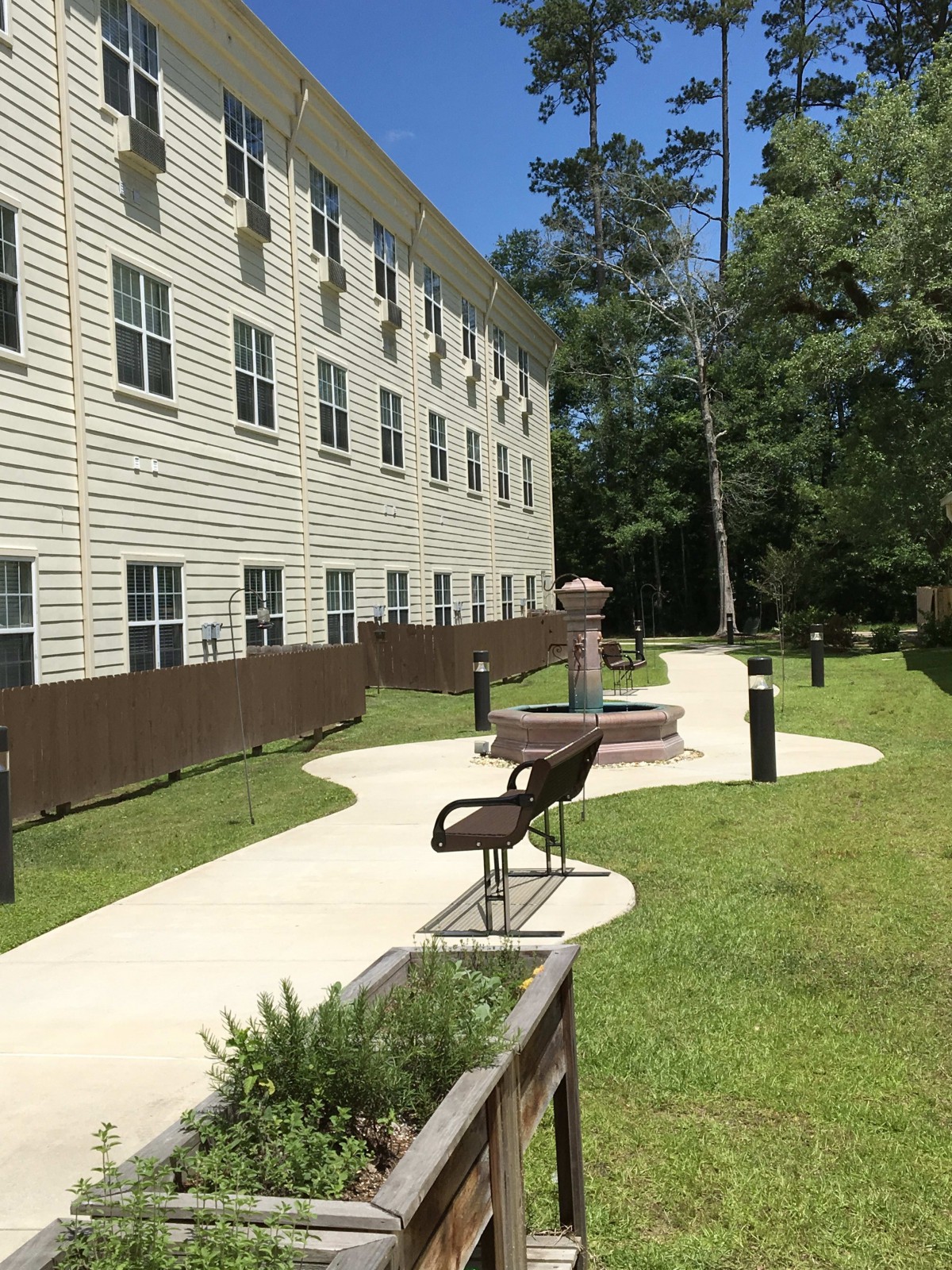 A pathway lined with benches next to a multi-story building, surrounded by trees and grass.