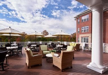 A spacious outdoor patio with comfortable seating and tables under umbrellas, adjacent to a multi-story building against a partly cloudy sky.