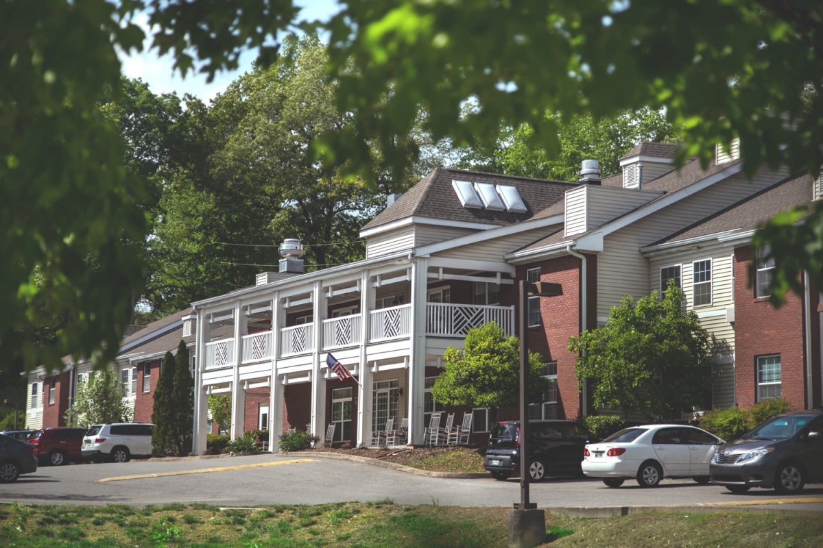 A two-story building with balconies surrounded by trees and parked cars.