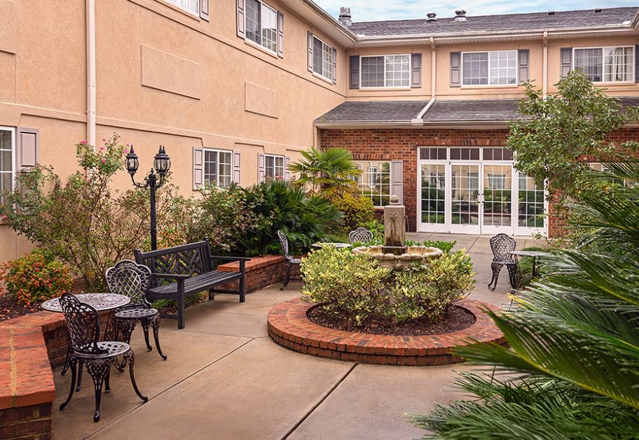 A charming courtyard featuring a fountain surrounded by benches and tables, with lush greenery and an inviting building exterior.