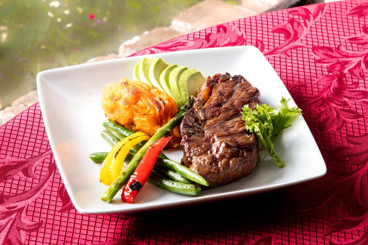 A gourmet dish featuring a perfectly cooked steak with grilled vegetables, sliced avocado, and a garnish on a stylish white plate.