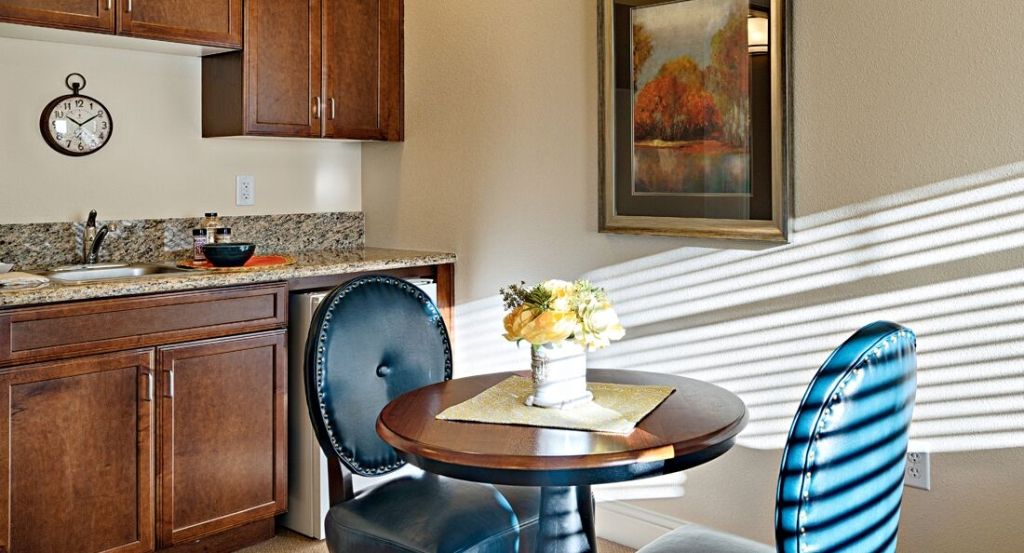 A cozy kitchen nook with a small round table, decorated with flowers and surrounded by elegant chairs, brightly lit by sunlight streaming through the window.