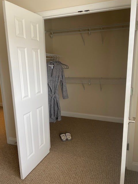 An empty closet with an open door, featuring a robe on hangers and slippers on the carpeted floor.