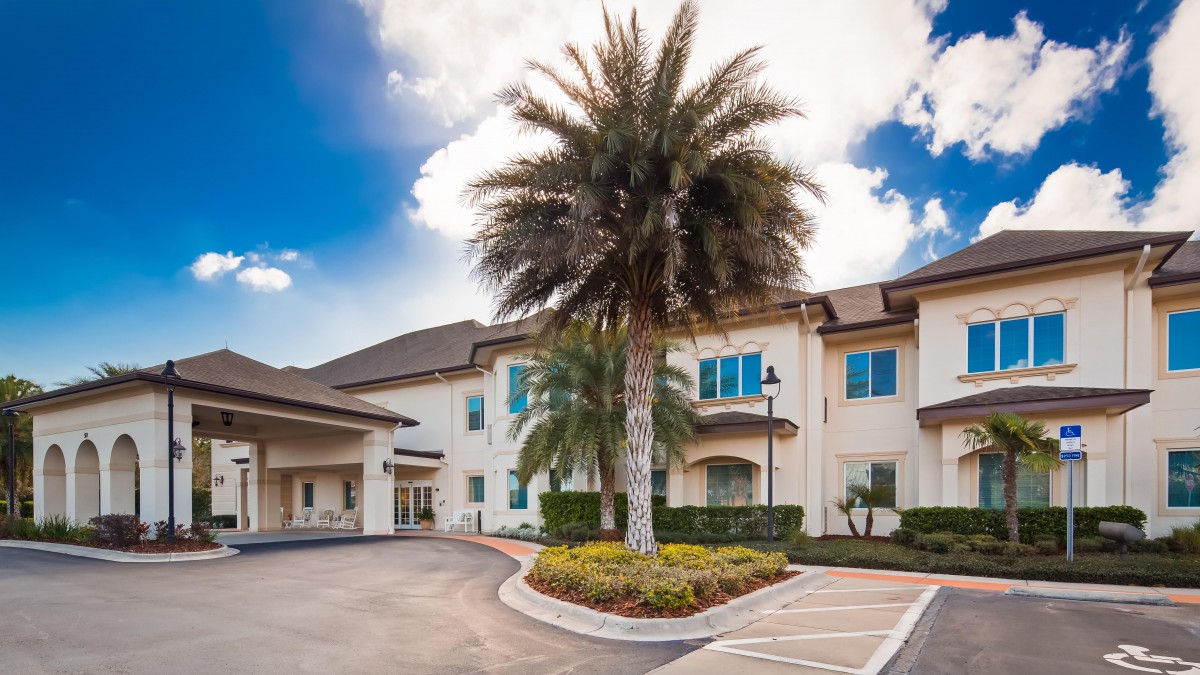 A modern building with a palm tree in the foreground and a clear blue sky with clouds.