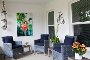 A cozy patio area featuring rattan chairs with navy cushions, vibrant artwork on the wall, decorative plants, and a small table for relaxation.
