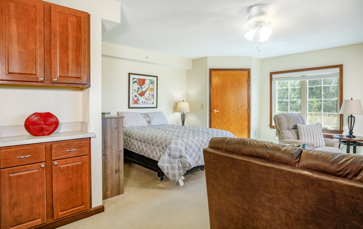 A cozy bedroom featuring a neatly made bed, artwork on the wall, and natural light streaming through large windows.
