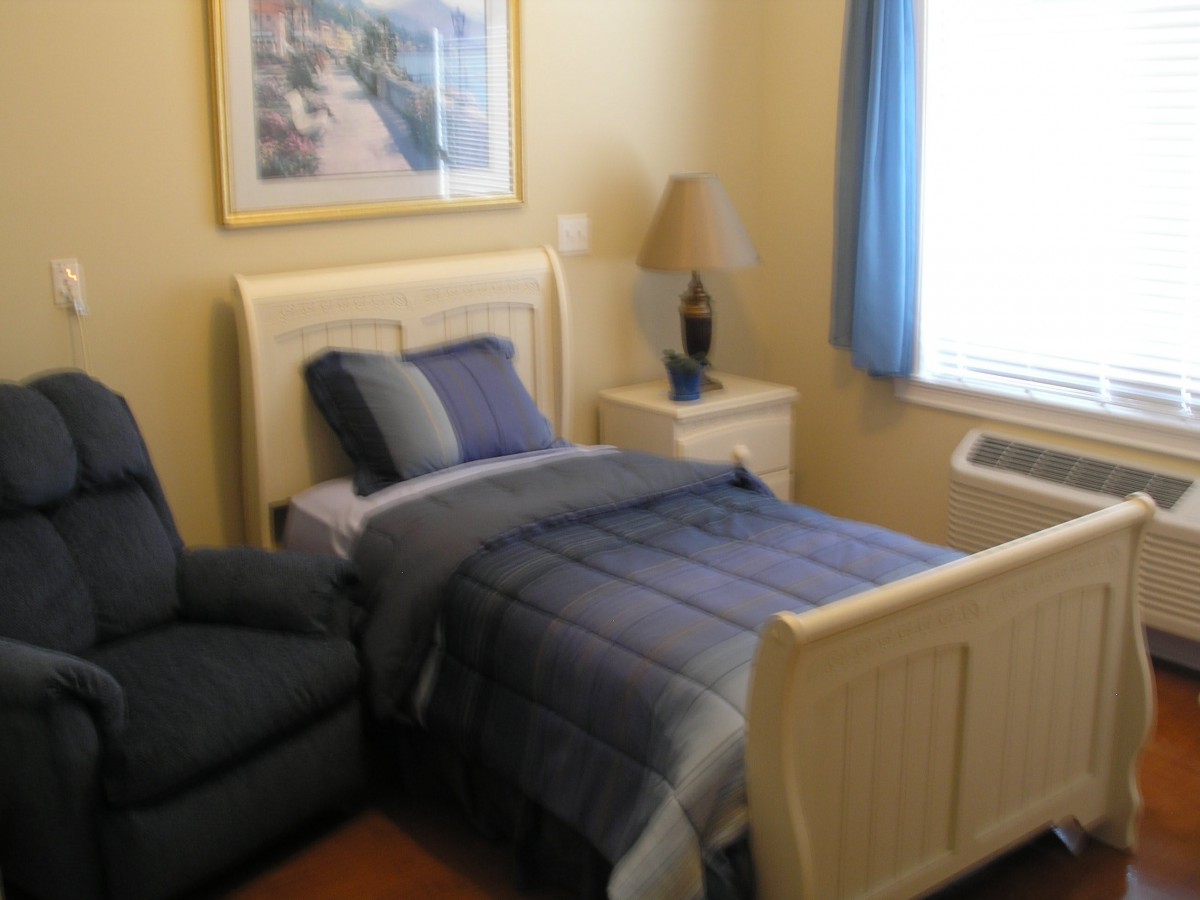 A cozy bedroom with a single bed featuring blue bedding, a nightstand with a lamp, and a window with blue curtains.