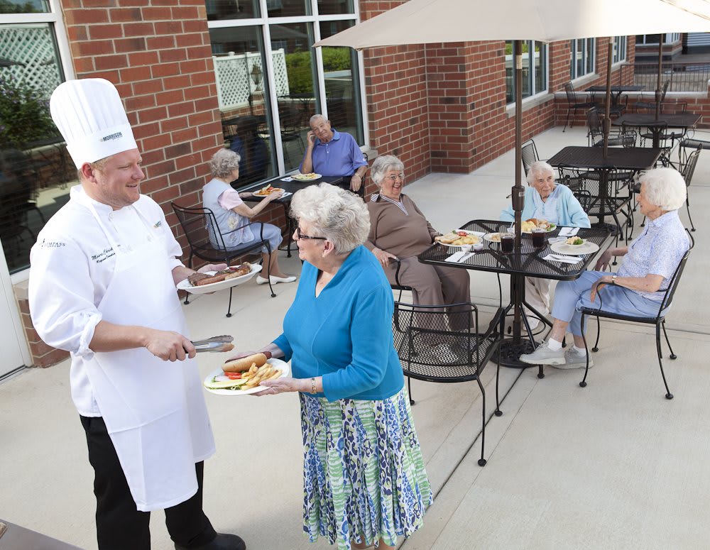 A chef serves food to a senior woman in an outdoor patio setting with other seniors enjoying the meal.