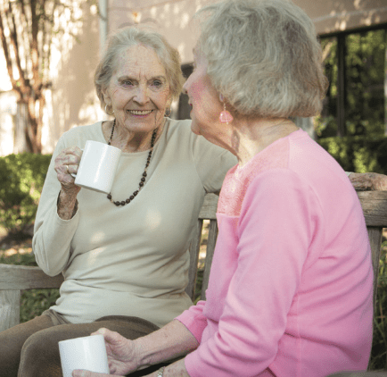 Two elderly women enjoying coffee and conversation on a bench outdoors.