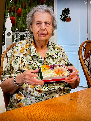 An elderly woman enjoys a moment at home, holding a colorful box of flowers.