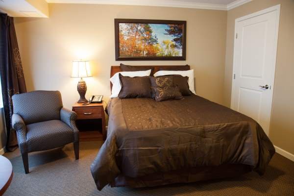 A cozy bedroom with a neatly made bed, accompanied by a bedside lamp and chair, featuring wall art above the headboard.