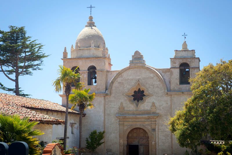 A picturesque church with Spanish colonial architecture, set against a clear blue sky and surrounded by palm trees.